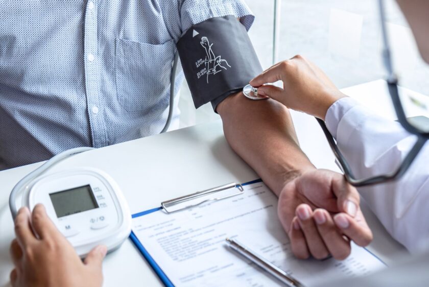 A medical professional, similar to Dr. Jai Bhagwan Dhull is using a manual blood pressure cuff and stethoscope to measure a patient's blood pressure, demonstrating a standard medical check-up. The patient's arm is resting on a table next to a digital blood pressure monitor and a clipboard with documents. highlighting a clinic setting for health assessment.