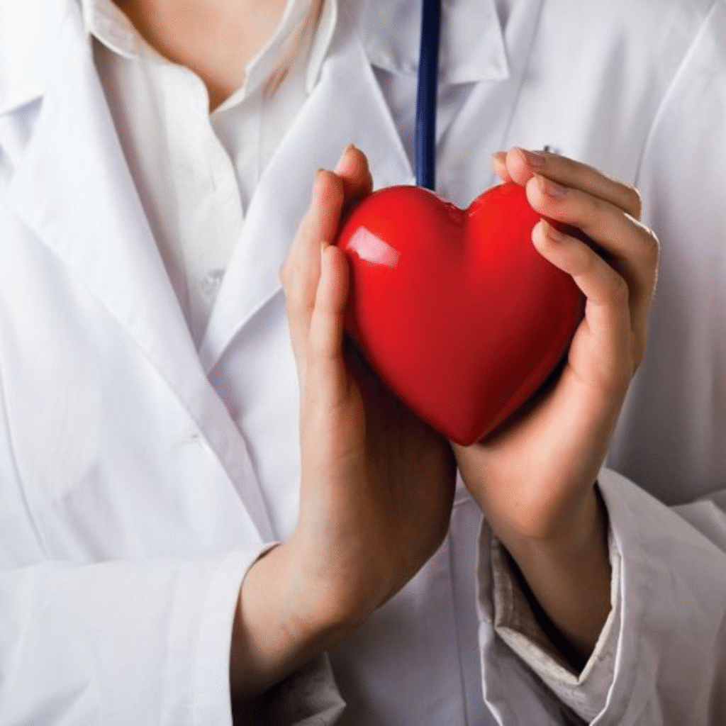 A medical professional in a white lab coat gently holding a red heart shaped model with both hands, symbolizing compassion, care and commitment to heart health and cardiology. This image conveys themes of patient centered care and cardiovascular wellness.