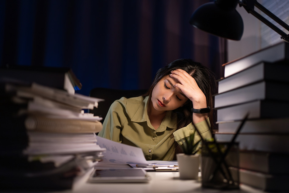 A stressed woman sits at a cluttered desk late at night, leaning forward with her forehead resting in one hand, surrounded by stacks of papers and books, illuminated by a single desk lamp—conveying fatigue and overwhelm.