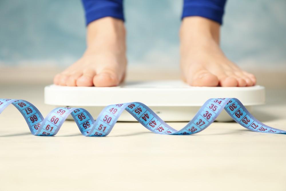 Close-up of a person standing barefoot on a weighing scale with a blue measuring tape in the foreground, symbolizing weight management and healthy lifestyle.