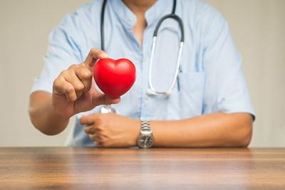 A doctor wearing a stethoscope holds a small red heart model in his hand, symbolizing heart health and cardiac care.

