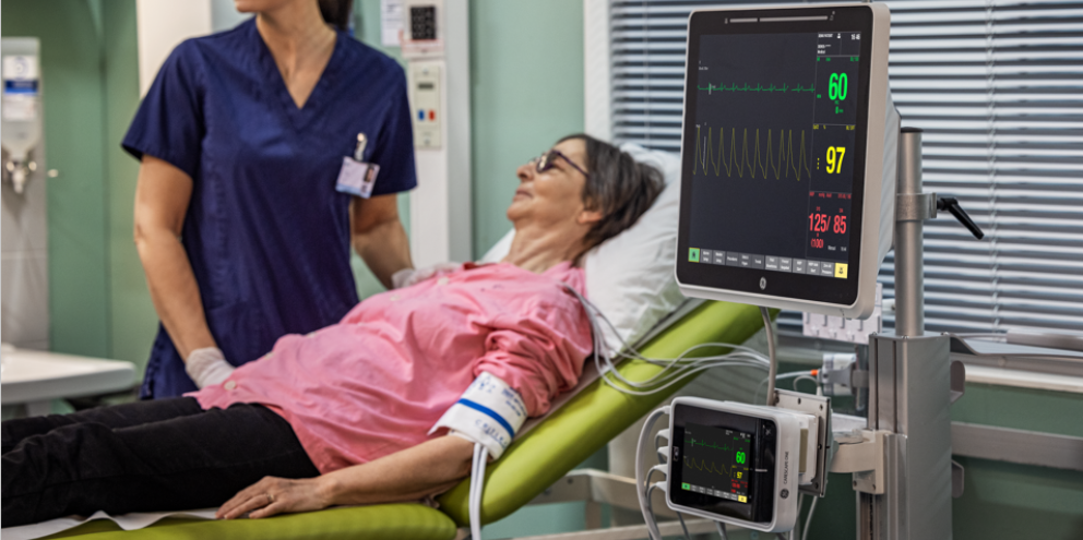 Elderly woman lying on a hospital bed being monitored by medical staff; vital signs displayed on a digital patient monitor showing heart rate, oxygen saturation, and blood pressure.