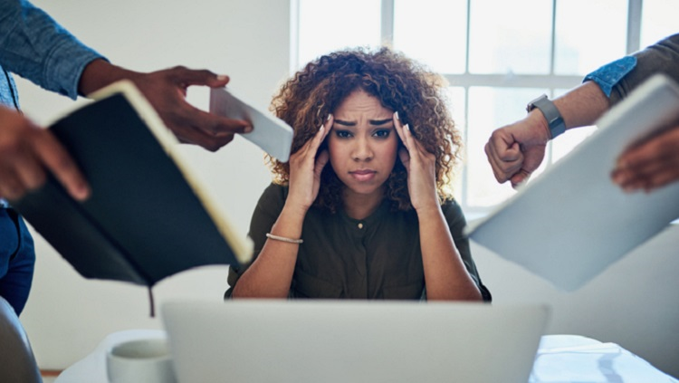 A stressed woman sitting in front of a laptop, holding her head while multiple hands around her demand attention with notebooks, papers, and devices — symbolizing workplace stress and pressure. This relates to stress management approaches discussed by Dr. Jai Bhagwan Dhull.