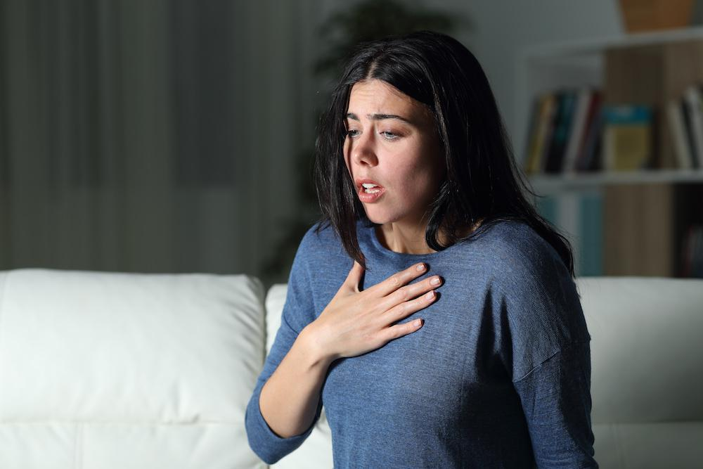 A young woman sitting on a sofa with a distressed expression, holding her chest as if experiencing shortness of breath or chest pain, representing symptoms of anxiety or heart issues, with health awareness guidance by Dr. Jai Bhagwan Dhull.