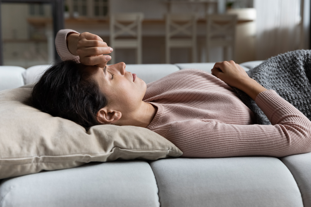 A woman lying on a couch with her hand on her forehead, appearing unwell or fatigued, symbolizing symptoms of illness or stress, with healthcare guidance by Dr. Jai Bhagwan Dhull.