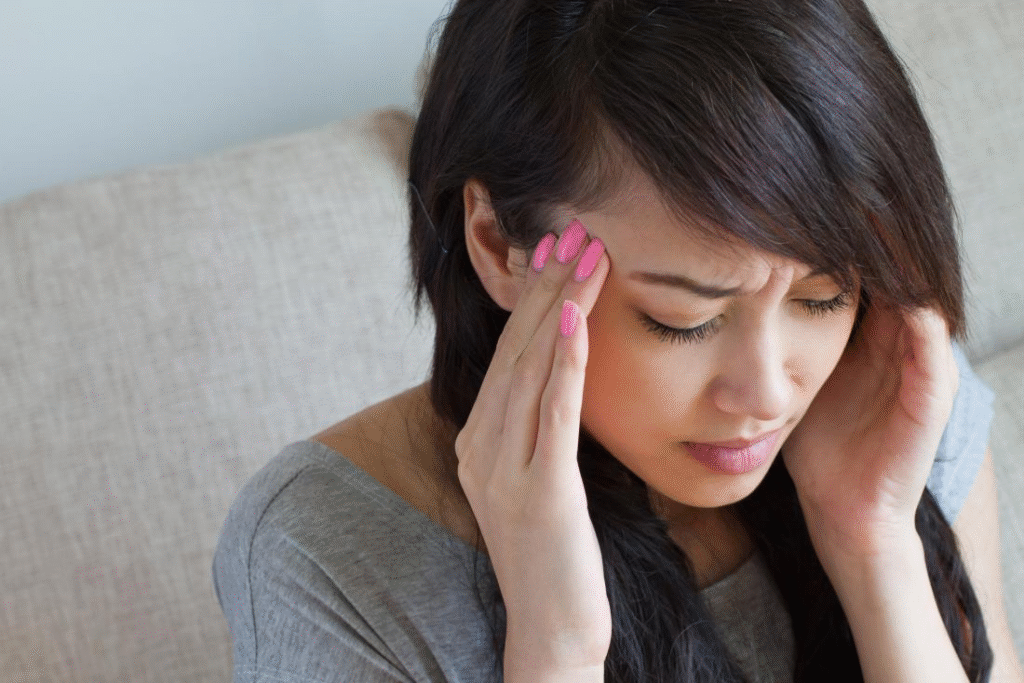 A young woman sitting on a sofa, holding her head with both hands and appearing to suffer from a severe headache or migraine, highlighting the importance of neurological and cardiovascular care with guidance from Dr. Jai Bhagwan Dhull.