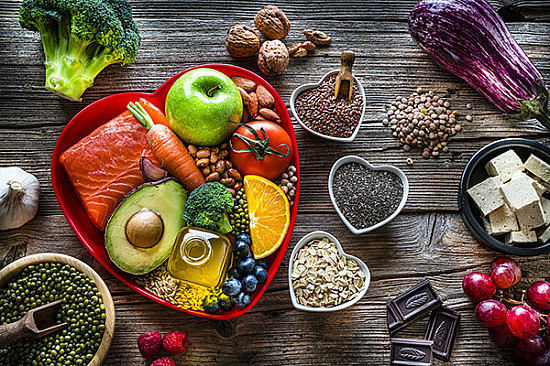 Assortment of healthy foods including salmon, avocado, fruits, vegetables, nuts, seeds, olive oil, and dark chocolate arranged in a heart-shaped dish on a rustic wooden table, symbolizing balanced nutrition and heart health.