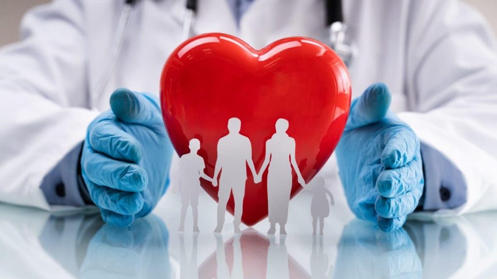 A doctor wearing blue gloves holds a large red heart with paper cutouts of a family—two adults and two children—standing in front of it, symbolizing healthcare and family heart protection.