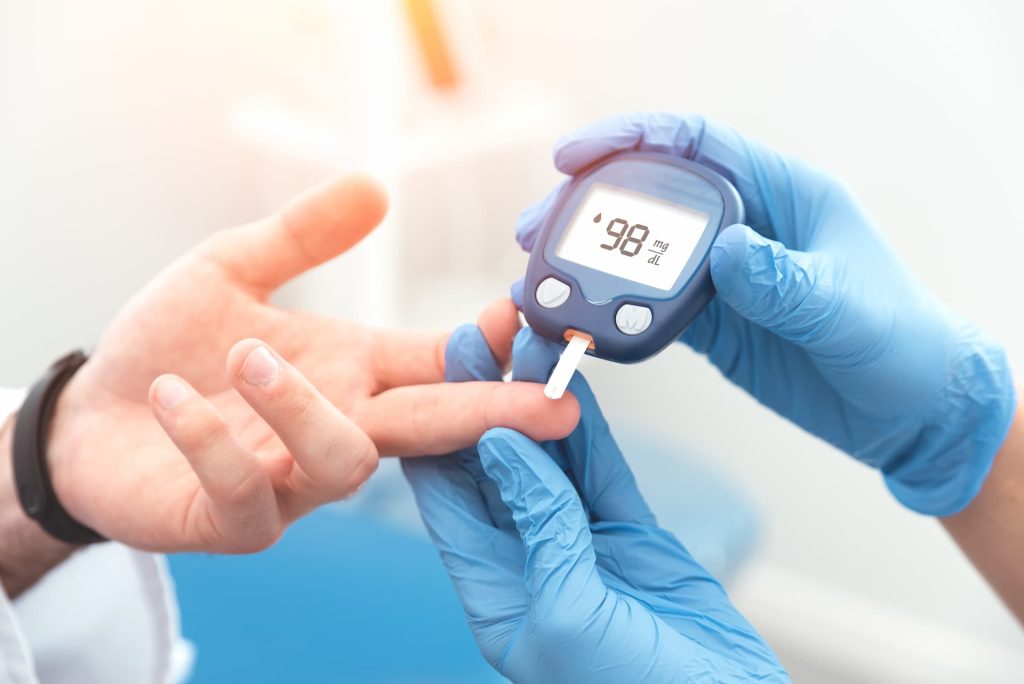 A healthcare professional wearing blue gloves checking a patient’s blood sugar level with a glucometer, showing 98 mg/dL on the screen — health awareness campaign by Dr. Jai Bhagwan Dhull.
