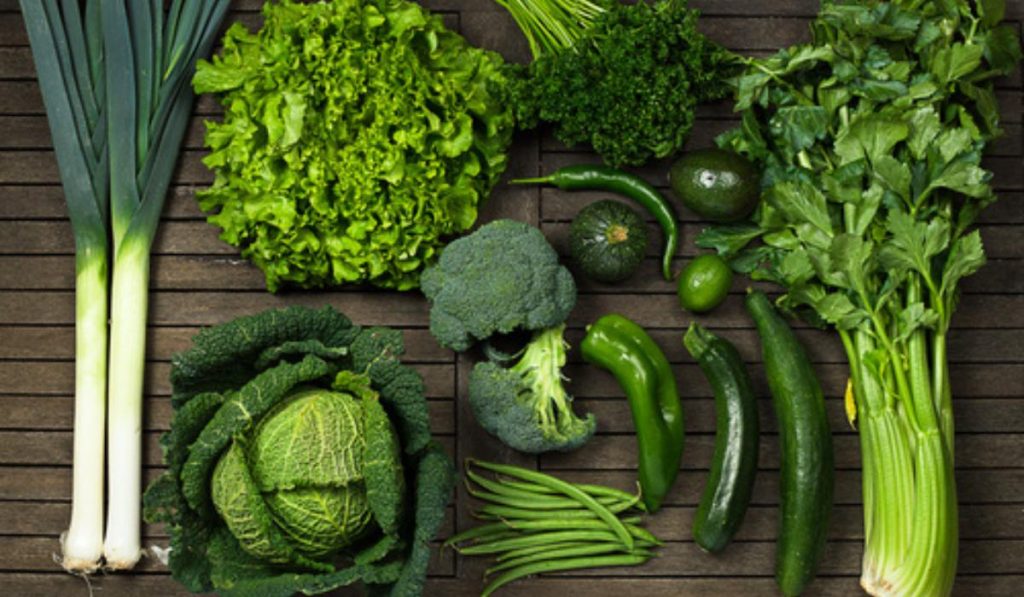 A variety of fresh green vegetables including broccoli, lettuce, cabbage, celery, cucumbers, leeks, green beans, parsley, and avocados arranged on a wooden surface — recommended by Dr. Jai Bhagwan Dhull for a healthy and balanced diet.