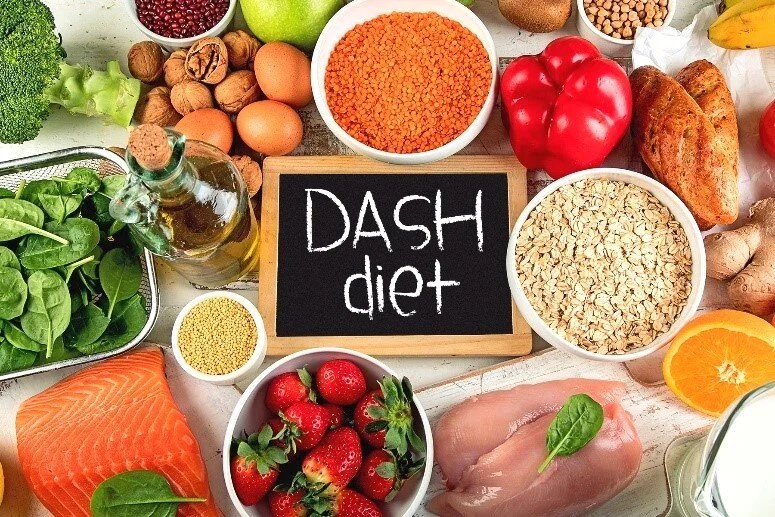 Healthy assortment of foods including salmon, chicken, eggs, spinach, lentils, oats, nuts, fruits, and vegetables surrounding a chalkboard with the text 'DASH diet' written on it.