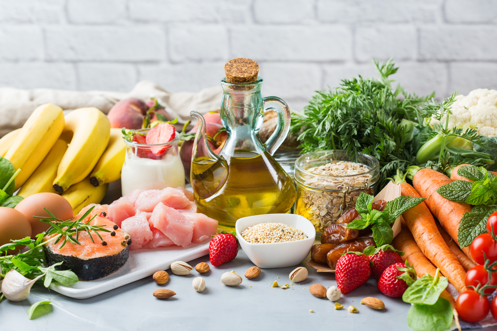 Assorted healthy foods including bananas, salmon, chicken, eggs, olive oil, oats, nuts, carrots, strawberries, and fresh vegetables displayed on a table, representing a balanced diet for overall wellness.