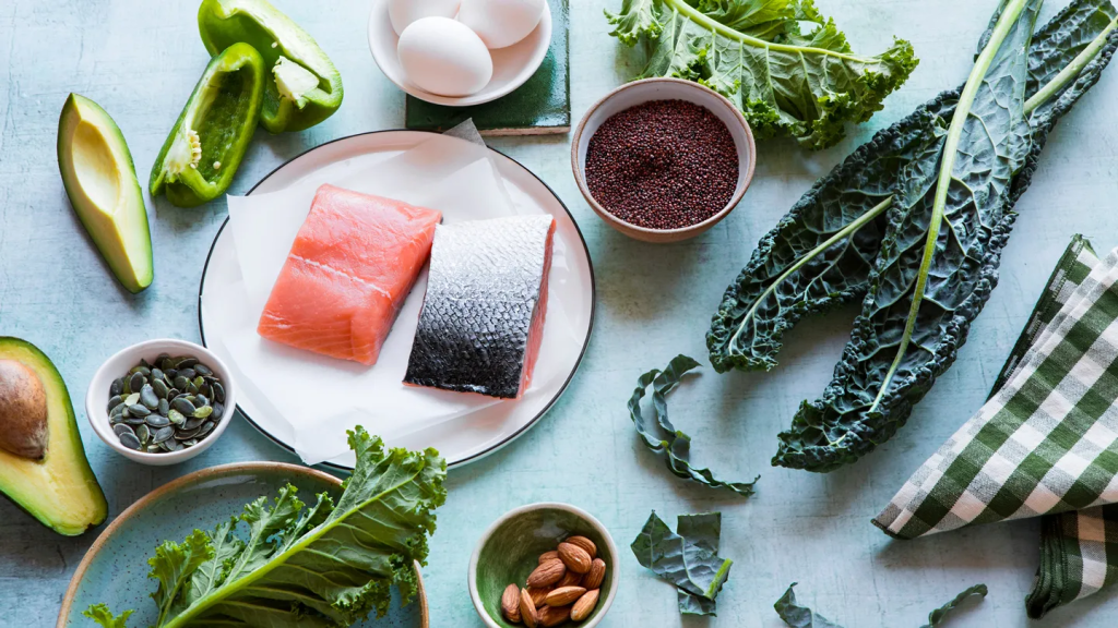 Fresh healthy foods including salmon fillets, avocado, kale leaves, green bell pepper, eggs, almonds, pumpkin seeds, and a bowl of grains arranged on a light blue surface.