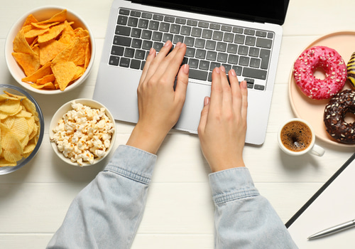 Person working on a laptop with hands on the keyboard, surrounded by bowls of chips, popcorn, coffee, and colorful donuts on a white desk.